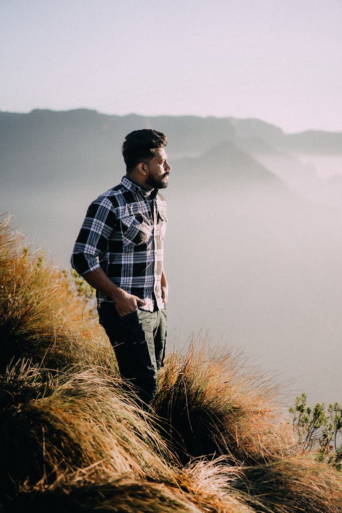 A man stands on a grassy hill with hands in pockets, gazing at a misty mountain view.