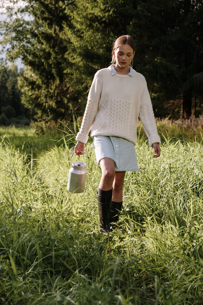 A woman in a sweater walks through a sunlit meadow carrying a can, surrounded by lush greenery.
