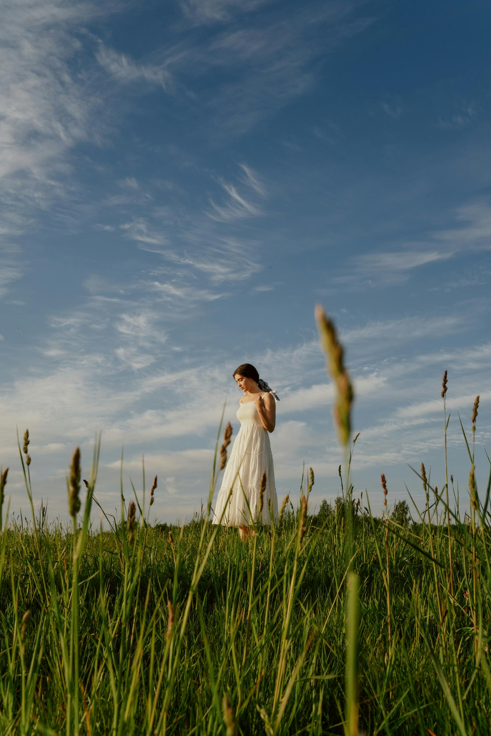 A woman in a white dress strolls through a grassy meadow under a clear sky.
