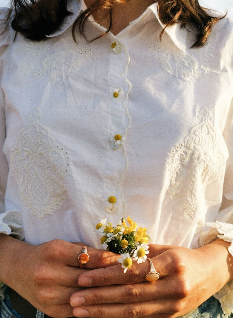 Close-up of a woman holding daisies, wearing an embroidered white shirt with a boho style.